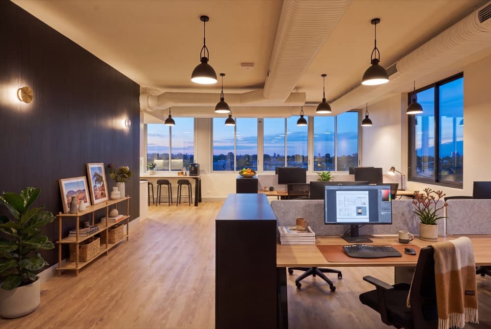An open plan office floor virtually staged at dusk with desks, seating, pendant lighting, shelving and planting